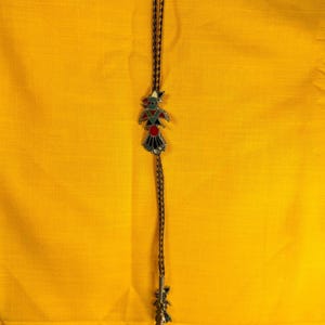 Puede incluir: Una corbata bolo con un colgante decorativo con un diseño colorido de un pájaro y formas geométricas. El colgante tiene detalles en rojo, verde y negro. La corbata está hecha de un cordón trenzado marrón oscuro.