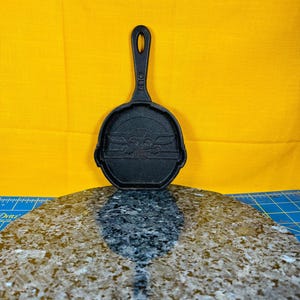 May include: A small, black cast iron skillet with a long handle and a circular cooking surface. The skillet features a raised image of a character from a popular science fiction franchise. The handle has a hole for hanging. The skillet is displayed against a yellow background.