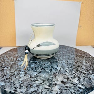 May include: A small, off-white ceramic vase with a gray rim and a gray and black feather design. The vase has a beaded tassel with tan feathers attached. The vase is on a speckled, gray and blue surface.