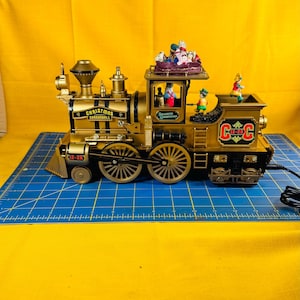 Może przedstawiać: Złoty świąteczny pociąg z napisem "Christmas Cannonball" i "C-C" z boku. Pociąg ma kilka małych figurek na górze i w oknach. Pociąg stoi na niebieskiej powierzchni kratki.