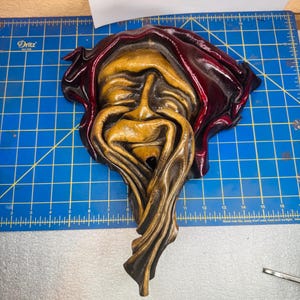 May include: A sculpted wall mask featuring a tan, flowing, face-like form draped in a deep burgundy hood. The mask is positioned on a blue cutting mat with visible white grid lines and inch markings, showing the object is approximately 10 inches wide.
