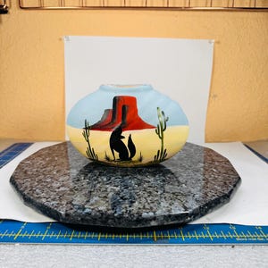 May include: Oval-shaped ceramic vase with a Southwestern desert landscape design. The vase features a red butte, cacti, and a black silhouette of two coyotes. The background is light blue and yellow.