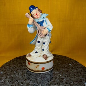 May include: A porcelain clown figurine playing a banjo. The clown is wearing a blue hat, a white and blue costume with black polka dots, and brown shoes. The figurine is on a round base with red and gold accents.