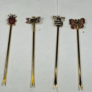May include: Four decorative cocktail picks with insect designs. The picks feature a ladybug, dragonfly, bee, and butterfly, each adorned with colorful rhinestones. The stems are gold-colored, and the tips are forked.