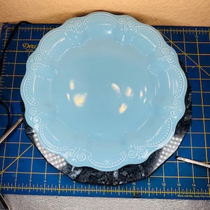 Puede incluir: Un plato de cerámica azul claro con borde festoneado y diseño floral en relieve. El plato es redondo y se asienta sobre una superficie gris oscuro. El diámetro del plato es de aproximadamente 25 cm.