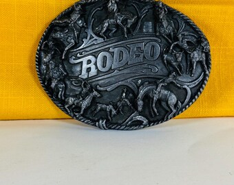 Puede incluir: Una hebilla de cinturón de rodeo de color plateado con un diseño detallado. La palabra "RODEO" se muestra de forma destacada en el centro, rodeada de representaciones de caballos y jinetes. La hebilla tiene un borde texturizado y está sobre un fondo amarillo.