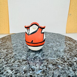 May include: A small, hand-painted ceramic figurine. The figurine is egg-shaped with a white base and a red and black design on the upper portion. The design includes a red vest-like shape with black accents. The figurine is on a speckled surface.