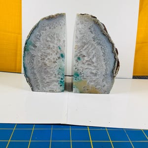 May include: Two agate bookends, each a semi-circular shape, with a gray and white interior and a dark brown outer edge. The bookends have areas of teal and yellow. The bookends are displayed on a white surface.