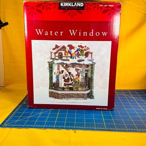 May include: A red box with the words "Water Window" and "Kirkland Signature" printed on it. The box contains a decorative water window scene with Santa Claus and other figures. The scene is set in a winter village.