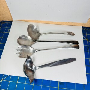 May include: A collection of four silver-toned metal serving utensils including two ladles, one large spoon, and one three-pronged serving fork. Each utensil features a smooth, polished finish with varying handle shapes and bowl designs.