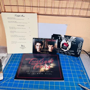May include: A collection of Twilight-themed memorabilia including a Twilight Menu, a Team Edward and Team Jacob New Moon card, a Twilight movie ticket, and a Twilight Tours illustrated guide book, all arranged on a blue cutting mat.