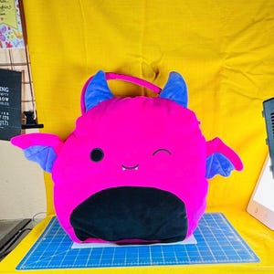 May include: A pink bat-shaped cushion with blue horns and wings. The cushion has a black belly and a smiling face with one winking eye. A pink handle is attached to the top.