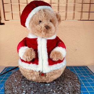 May include: A teddy bear dressed in a red Santa suit with white trim and a matching hat. The bear is light brown and has a button on the front of the suit. The bear is sitting on a dark grey stone.