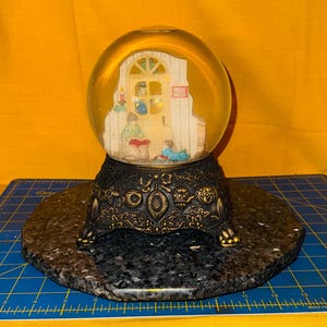 May include: A snow globe with a gold-colored base and a black decorative stand. Inside the globe, a miniature scene depicts figures in a room. The base has an ornate design. The globe sits on a dark gray, hexagonal base.