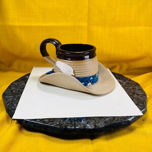May include: A ceramic mug with a brown interior and handle, resting on a tan cowboy hat with a blue patterned band. A white feather is attached to the hat. The mug and hat are on a white surface, set against a yellow background.