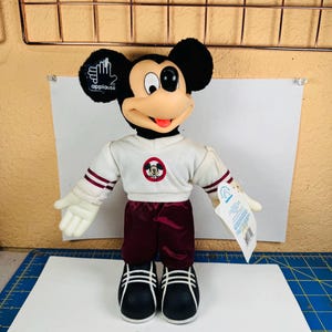 May include: A plush Mickey Mouse doll featuring a cream-coloured long-sleeve shirt with maroon stripes on the sleeves and a circular graphic on the chest, paired with maroon trousers and black shoes with white stripes. The doll has a plastic face and ears.