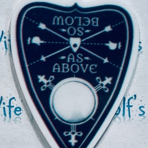 May include: A blue and white planchette with the text "Below As Above" and a circle in the centre. The planchette is a triangular shape with a pointed top.