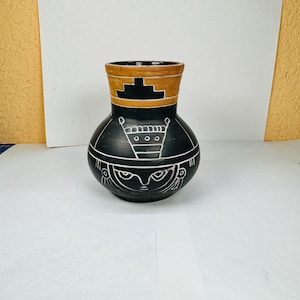 Puede incluir: Un jarrón de cerámica negro con cuerpo redondeado y borde marrón. El jarrón presenta diseños geométricos blancos, incluido un motivo similar a una cara y una serie de formas alrededor del cuello. El jarrón es una pieza decorativa.