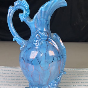 May include: A blue ceramic pitcher with a swirling, marbled design and gold accents. The pitcher has a wide, round base and a tall, slender spout.