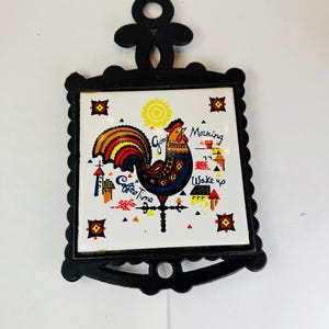 May include: Black metal trivet with a ceramic tile insert. The tile features a colorful rooster illustration with the words "Good Morning, Coffee Time, Wake Up". The trivet has a decorative scalloped edge and a handle.