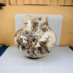 Puede incluir: Un jarrón de cerámica color crema con cuerpo redondeado y boca ancha. El jarrón presenta un patrón único de rayos negros y formas geométricas blancas. La cerámica hecha a mano es un artículo decorativo.