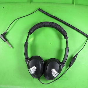May include: Black over-ear headphones with silver accents on the earcups and a padded headband. The headphones have a coiled cord and a 3.5mm audio jack. The headphones are on a green surface.
