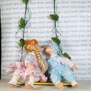 May include: Two porcelain dolls sitting on a yellow swing. The girl doll is wearing a pink and white dress with a pink bow in her hair. The boy doll is wearing a blue and white overall with a blue hat. The dolls are looking at each other.