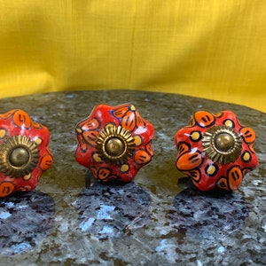May include: Three ceramic drawer knobs with a floral design. The knobs are red with orange and yellow accents and have a gold metal center.