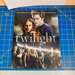 Puede incluir: Un libro de tapa blanda titulado Twilight: The Complete Illustrated Movie Companion de Mark Cotta Vaz. La portada presenta una gran fotografía de dos personajes principales sobre un grupo de personajes secundarios bajo un cielo nublado.