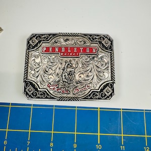 Peut inclure: Une boucle de ceinture rectangulaire en métal argenté avec des gravures à volutes complexes, une figure centrale de cheval au galop et des accents en émail rouge avec le texte PENDLETON WHISKY et Let'er Buck. Elle mesure environ 9 cm de large.