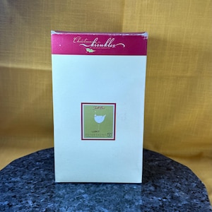 Op de afbeelding: Een witte doos met een rode bovenkant en een groene en witte illustratie van een vogel aan de voorkant. De doos heeft de tekst "Christmas Krinkles" op de bovenkant en "Tall Bird" aan de voorkant. De doos heeft ook de tekst "Department 56" aan de voorkant.