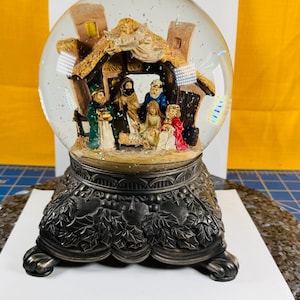 May include: A snow globe featuring a Nativity scene with figures inside a clear glass sphere. The base is a dark, ornate design. The scene includes figures in colorful robes, a stable, and glitter. The globe sits on a decorative base.