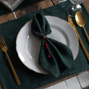 Puede incluir: Una mesa rústica con un camino de mesa y manteles individuales de lino verde oscuro. Una hogaza de pan descansa sobre una bandeja de madera con un cuchillo. Un plato blanco con una servilleta verde y cubiertos dorados está listo para una comida. Un vaso de agua y romero.