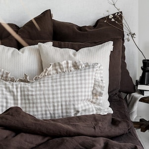 Pode incluir: Uma cama com roupa de cama castanha escura, incluindo um edredom e almofadas. As almofadas são de vários estilos, incluindo branco texturizado e um padrão xadrez vichy com uma borda de babados. Uma revista está aberta na cama.