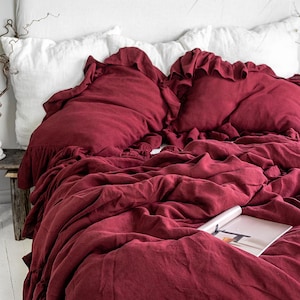 May include: A bed with a white headboard and white pillows. The bed is covered with a deep red duvet and matching ruffled pillowcases. A wooden side table holds decorative items.