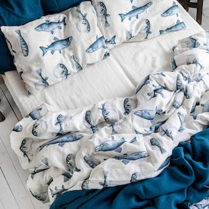 Peut inclure: Un lit avec des draps blancs, des oreillers bleus et une housse de couette avec un motif de poissons bleus. Une couverture bleue unie est drapée au pied du lit. La pièce a des murs blancs et un sol en bois.