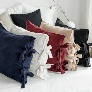 Puede incluir: Una colección de cojines de lino en varios colores, incluyendo azul marino, blanco, burdeos y beige. Cada cojín rectangular presenta un cierre de lazo. Los cojines están dispuestos sobre una cama blanca, creando una estética acogedora y elegante. Aproximadamente 40 cm x 60 cm.