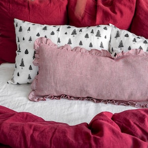 Op de afbeelding: Een bed met een witte hoofdbord en beddengoed in rood- en wittinten. Het bed is opgemaakt met verschillende rode kussens, een wit kussen met een zwart boompatroon en een rood-wit geruit kussen met een gerimpelde rand. Een rood dekbed bedekt het bed.