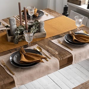 May include: A rustic wooden dining table set for a meal. The table is adorned with place settings featuring dark gray plates, gold-colored cutlery, and folded mustard-colored napkins. A mustard-colored table runner and beige placemats add warmth.