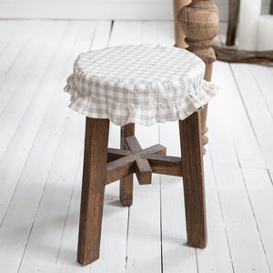 May include: A wooden stool with a round, gingham-patterned seat cover and a frilled edge. The stool has four sturdy legs and a cross-shaped support. The background includes a wooden table and a white-painted floor.