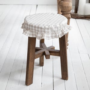 Pu&ograve; includere: Sgabello in legno con rivestimento a quadretti e finiture a balze. Lo sgabello ha una base a quattro gambe ed &egrave; realizzato in legno scuro. Il rivestimento della seduta presenta un motivo a quadretti beige chiaro e bianco. Lo sgabello &egrave; in una stanza con un tavolo di legno.