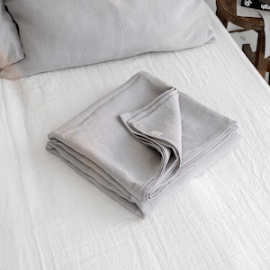 Puede incluir: Una cama de lino blanco con una manta de lino gris doblada en la parte superior. Dos almohadas de lino gris están en la cama. La cama está hecha y la manta está doblada cuidadosamente.