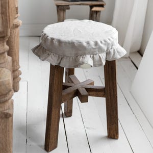 Pu&ograve; includere: Uno sgabello in legno con una seduta rotonda in lino chiaro con un bordo a balze. Lo sgabello ha una struttura in legno rustico marrone scuro con un supporto a forma di croce. Lo sfondo comprende un tavolino in legno e assi del pavimento verniciate di bianco.