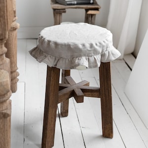 May include: A wooden stool with a round, light-coloured linen seat cover with a ruffled edge. The stool has a dark brown, sturdy wooden frame with a cross-shaped support. The background includes a small wooden table and white painted floorboards.