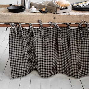 May include: A wooden table with a black and white gingham valance. The table is set with a cast iron pan, a metal jug, and a wooden tray holding bread. Black plates and cutlery are stacked on the right.