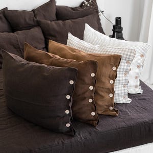 May include: A bed with dark brown linen bedding and several decorative pillows. The pillows are in shades of brown and tan, with button detailing. The bed is made up with a white headboard and a dark brown fitted sheet.