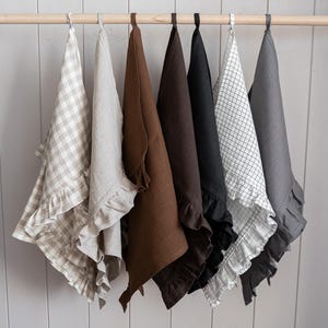 May include: A selection of ruffled linen tea towels in a range of colours, including beige gingham, natural, brown, black, and grey. The towels are suspended from a wooden rail with metal hooks, displaying a farmhouse or cottage aesthetic.