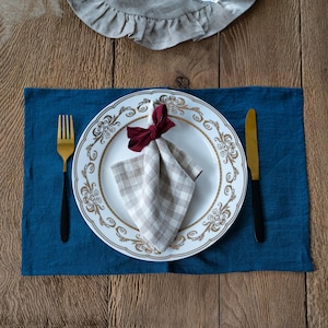 May include: A formal table setting featuring a blue linen placemat, a white plate with gold detailing, and a checkered napkin tied with a burgundy bow. A gold fork and knife are included. The table is made of wood.