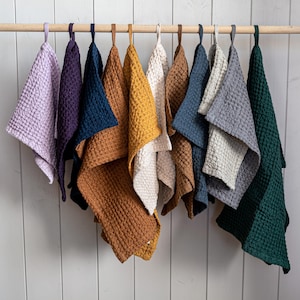 Puede incluir: Una colección de toallas de mano con tejido de gofre en varios colores, incluyendo lavanda, morado, azul marino, mostaza y verde, colgadas de una barra de madera. Las toallas tienen una superficie texturizada y se exhiben sobre un fondo de madera pintada de blanco.