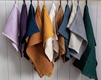 Puede incluir: Una colección de toallas de mano con tejido de gofre en varios colores, incluyendo lavanda, morado, azul marino, mostaza y verde, colgadas de una barra de madera. Las toallas tienen una superficie texturizada y se exhiben sobre un fondo de madera pintada de blanco.
